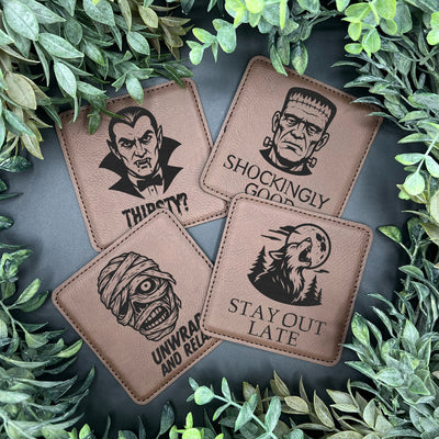 Four brown leather coasters with Halloween-themed designs on a dark surface surrounded by green leaves.