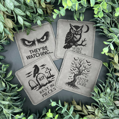 Four coasters with spooky designs on a dark surface surrounded by green leaves.
